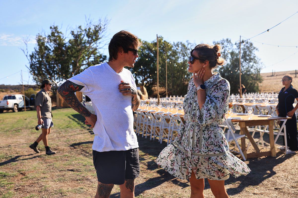 Tiffany and an event organiser discussing logistics at an outdoor event setup with decorated tables and string lights in the background.