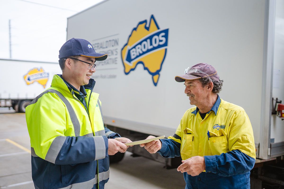 Two workers in high-visibility uniforms exchanging documents in front of a Brolos-branded delivery truck.