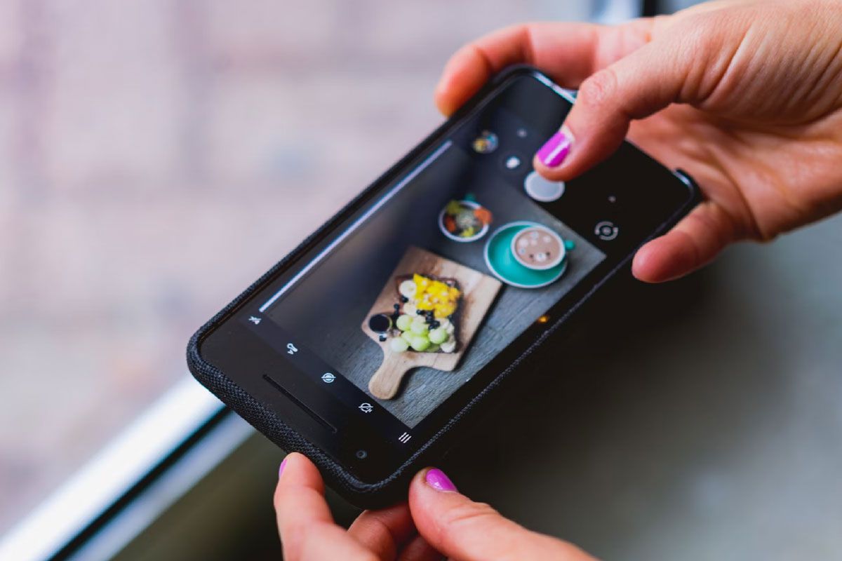 Close-up of a smartphone capturing a photo of a food platter with colourful fruit and desserts.